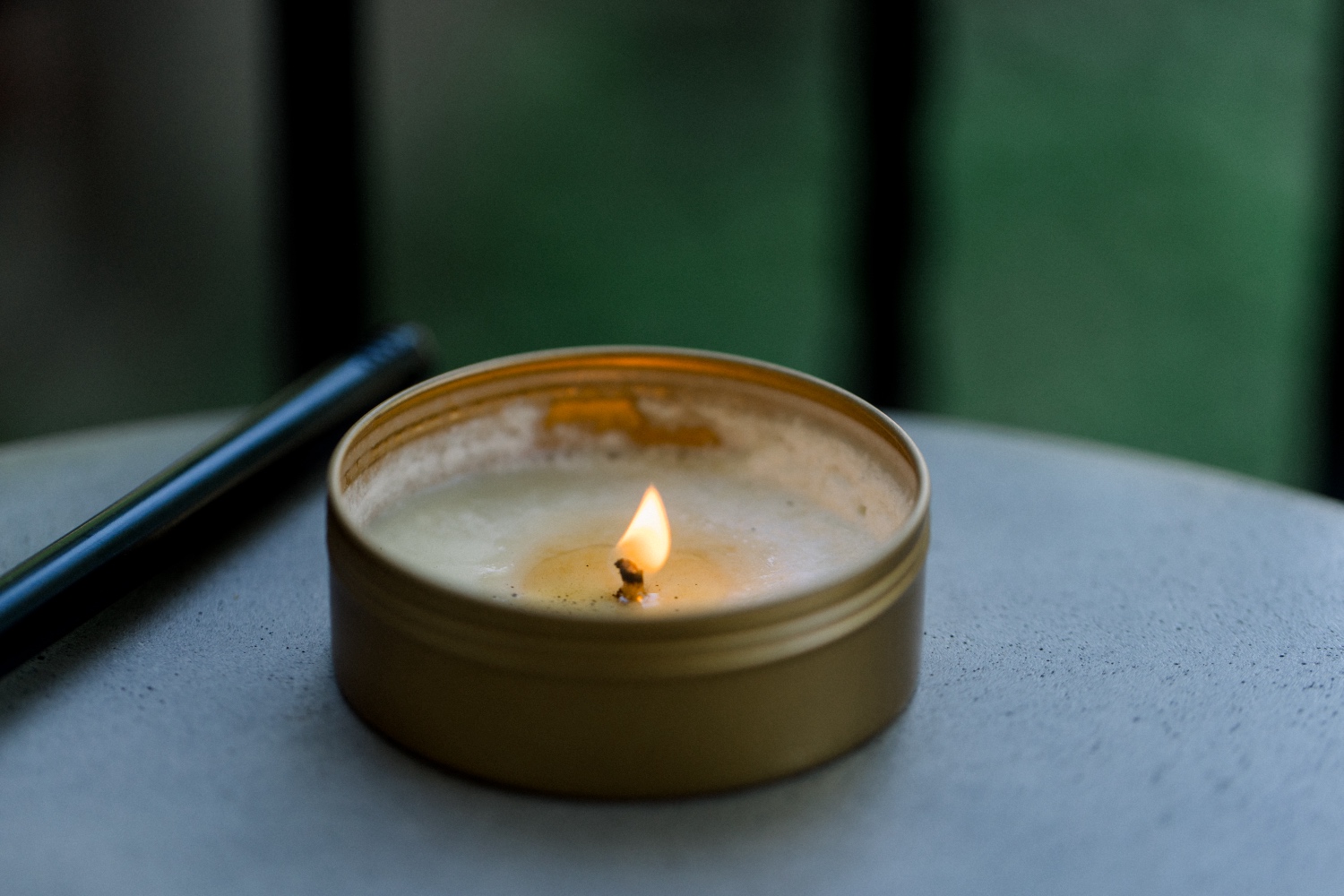A close-up photo of a lit citronella candle sitting on a concrete table with a long lighter lying next to the candle. The black iron bars of a porch and green grass are blurred in the background.