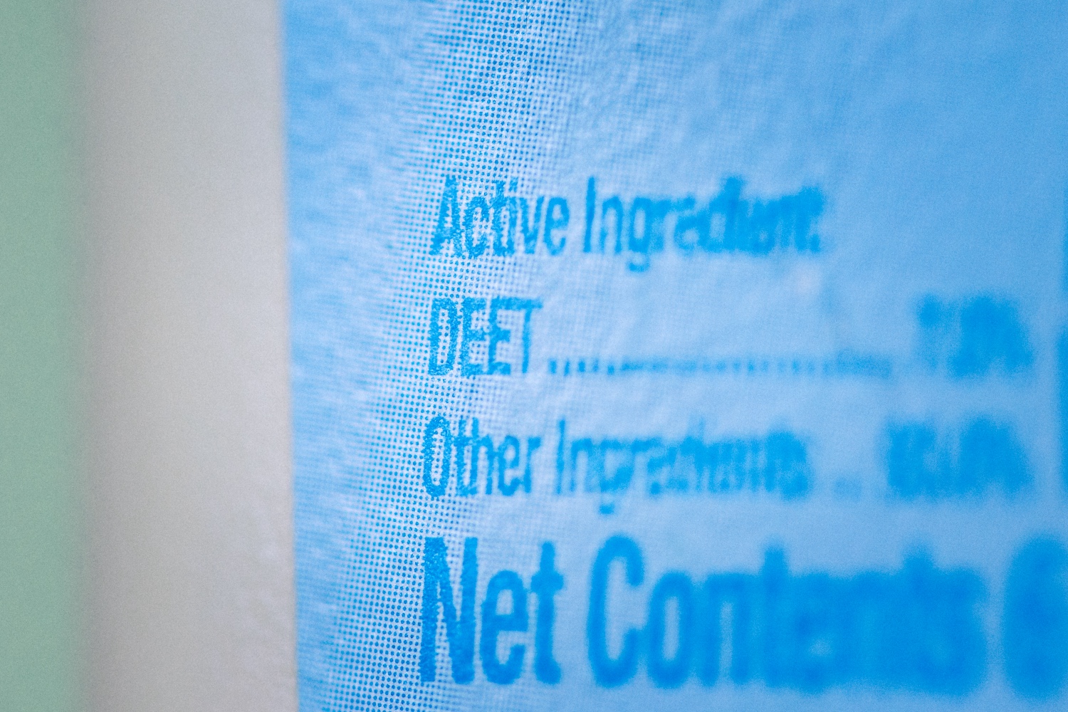 A macro photo of the list of active ingredients in a mosquito repellent. The label is light blue with darker blue writing on a white bottle, and the text reads: 